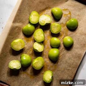 Halved green tomatillos arranged on a parchment-lined baking sheet, glistening with olive oil, prepared for oven roasting.
