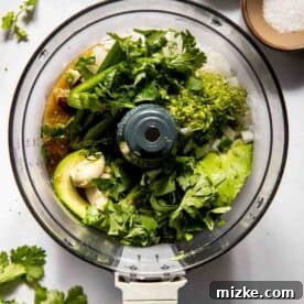 Overhead view of a food processor filled with fresh avocado, cilantro, lime zest, onions, and other ingredients, with bowls of salt and pepper nearby on a white surface, ready for blending into a vibrant salsa.