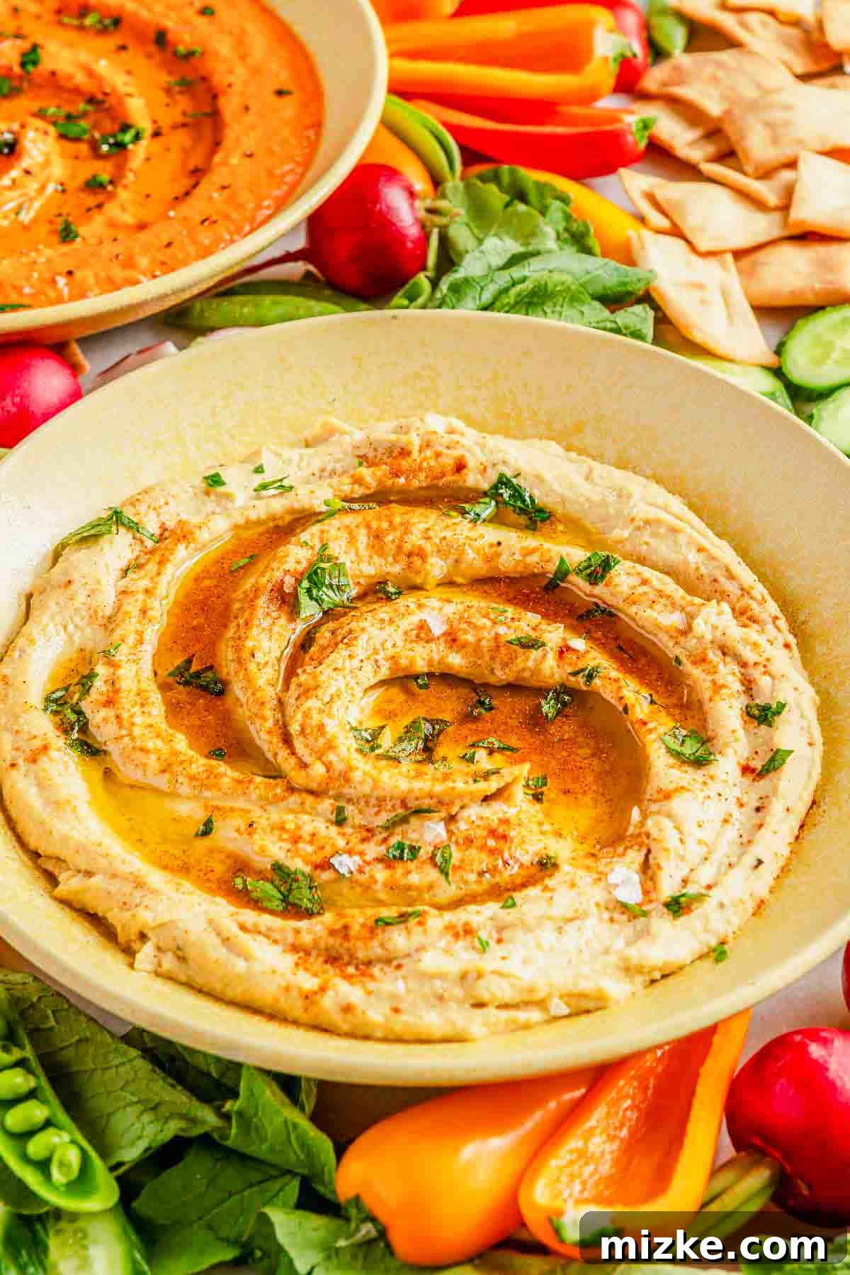 Robust Garlic Hummus 2 A beautifully presented bowl of garlic hummus, generously drizzled with golden olive oil, sprinkled with fresh herbs like parsley, and a dash of paprika. It's surrounded by an inviting array of colorful, fresh vegetables such as cucumber slices, carrot sticks, cherry tomatoes, and warm pita bread, inviting a delightful dip.