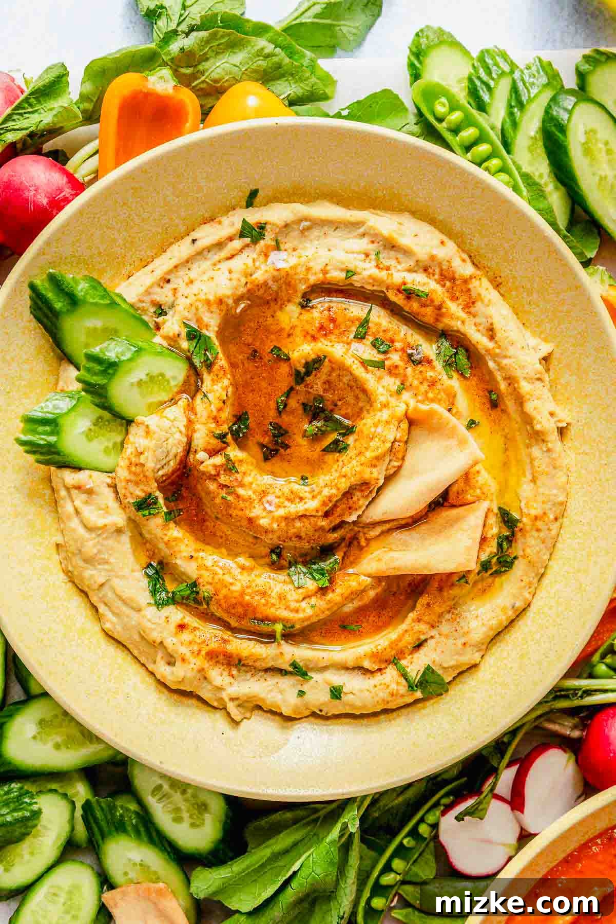 Robust Garlic Hummus 3 An overhead shot of a rustic wooden board featuring a generous bowl of garlic hummus, elegantly garnished with a swirl of olive oil, a sprinkle of vibrant paprika, and a scattering of fresh chopped parsley. The board is laden with an assortment of accompaniments including crunchy cucumber slices, warm pita bread wedges, and various fresh vegetables, creating an inviting spread for a healthy snack or appetizer.