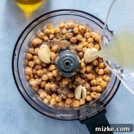 A food processor bowl filled with chickpeas, garlic, spices, and a lemon juice being poured in, with a bottle of olive oil nearby on a light blue surface.