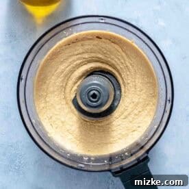 Overhead view of a food processor filled with smooth, blended hummus next to a bottle of olive oil on a light surface.