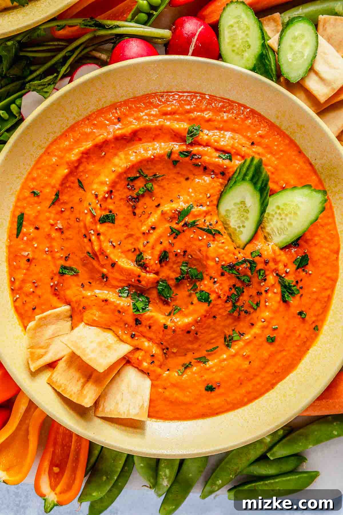 Fire-Roasted Red Pepper Dip 3 A vibrant bowl of roasted red pepper hummus, artfully garnished with chopped herbs and cucumber slices, perfectly complemented by a selection of crispy pita chips and various fresh vegetables.
