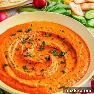 A bowl of red pepper hummus is garnished with herbs and surrounded by fresh vegetables and pita chips.
