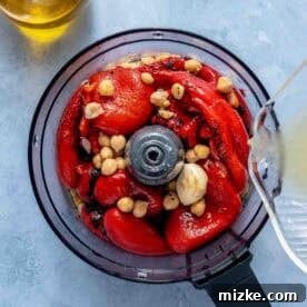 Fire-Roasted Red Pepper Dip 5 A food processor bowl filled with roasted red peppers, chickpeas, and garlic, with liquid being poured in; a jar of olive oil is nearby.