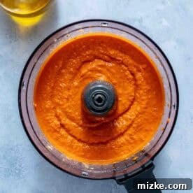 Fire-Roasted Red Pepper Dip 6 A food processor filled with blended red pepper hummus sits on a light gray surface next to a glass container of olive oil.