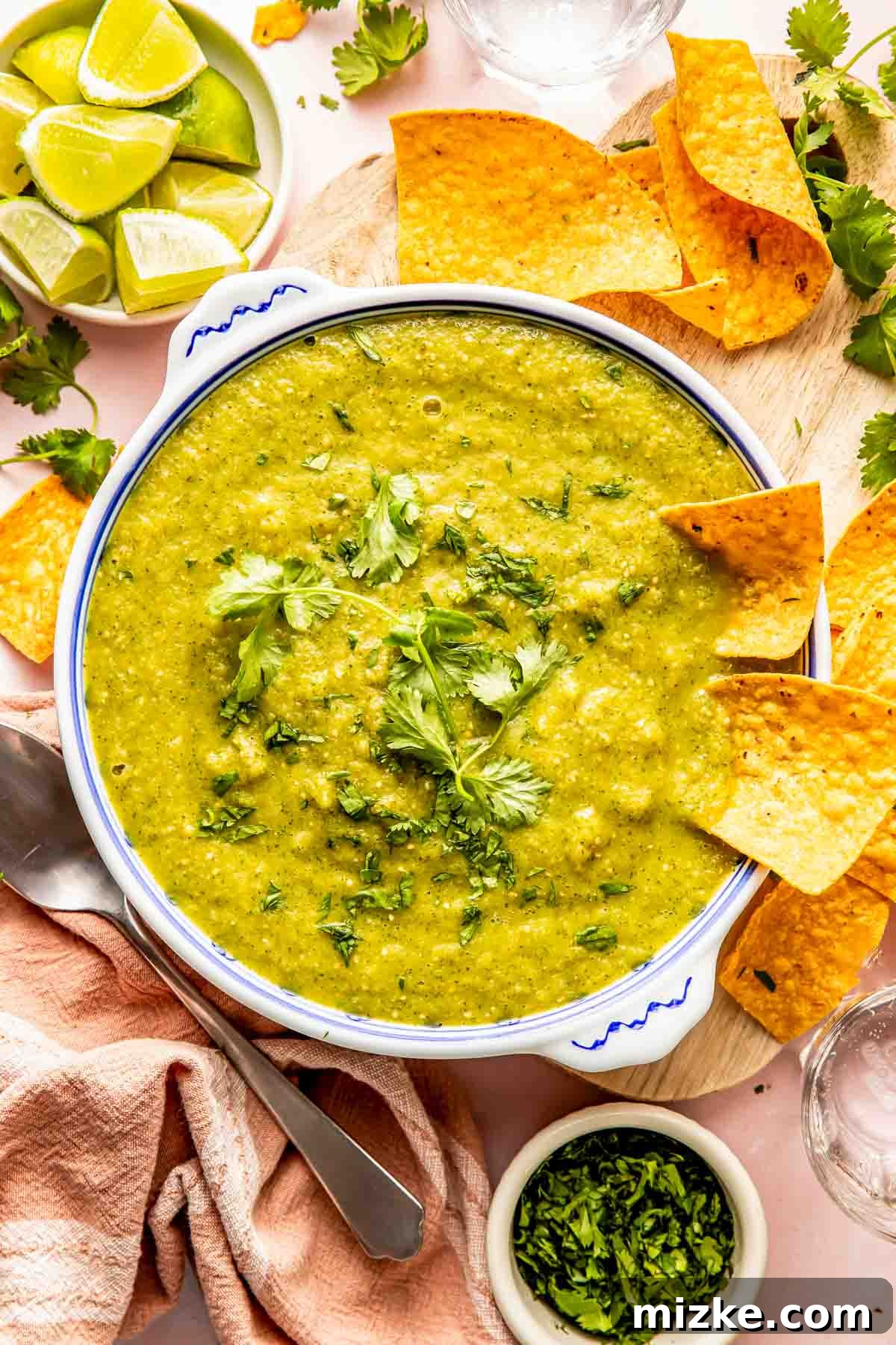 Fire-Roasted Salsa Verde 2 A bowl of green salsa garnished with cilantro, surrounded by tortilla chips, lime wedges, and a small bowl of chopped cilantro on a wooden board.