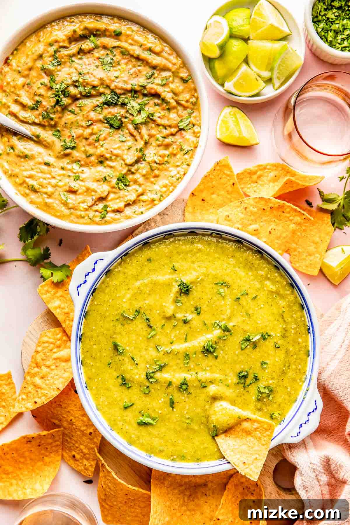 Fire-Roasted Salsa Verde 4 Two bowls of green and yellow dips garnished with herbs, surrounded by tortilla chips, lime wedges, and fresh cilantro on a white surface.