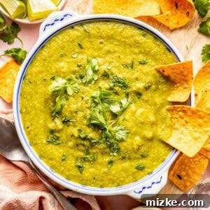 A bowl of green salsa garnished with cilantro, surrounded by tortilla chips, lime wedges, and a small bowl of chopped cilantro on a wooden board.