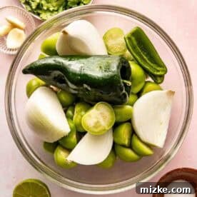 A glass bowl filled with tomatillos, halved onions, a poblano pepper, and a jalapeño. Surrounding the bowl are cilantro, garlic cloves, lime halves, and a small bowl of salt.