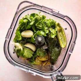 A blender filled with fresh cilantro, garlic cloves, jalapeño, and other green ingredients, viewed from above.