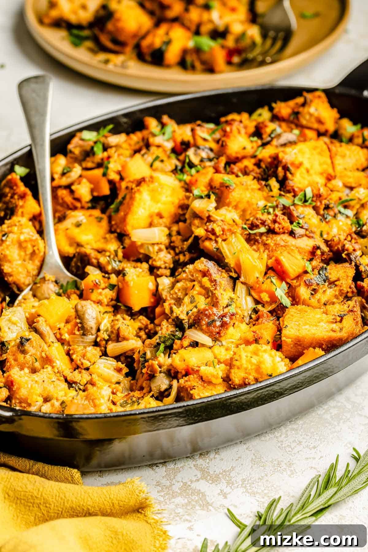 Fiery Chorizo Cornbread Bake 4 a metal spoon lies on the side of the chorizo cornbread stuffing in the oval baking dish next to fresh rosemary and a yellow tea towel