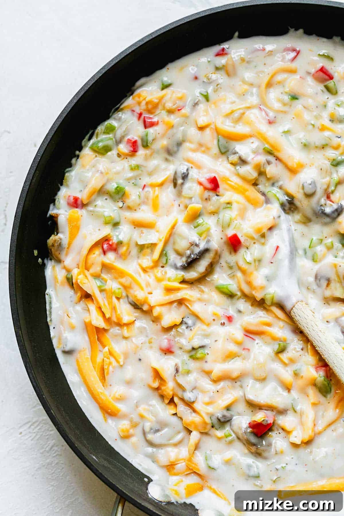 Cheesy Spaghetti Bake 4 Vegetable mixture combined with milk, cream of mushroom soup, and shredded cheese in a skillet, being stirred with a wooden spoon.