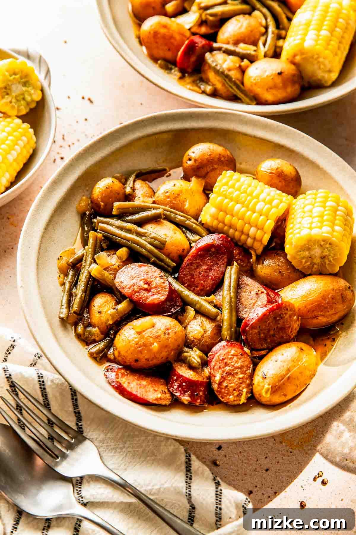 Slow Cooker Bayou Blend Potatoes 3 A beautifully arranged bowl showcasing slices of savory sausage, tender baby potatoes, crisp green beans, and a side of corn on the cob, all presented with a neatly folded napkin and forks.