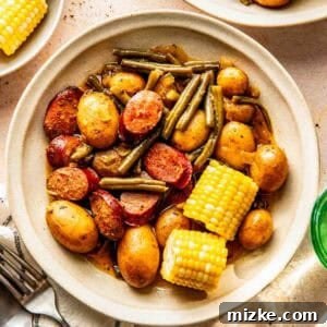 A bowl containing tender baby potatoes, vibrant green beans, savory sausage slices, and sweet corn on the cob, served elegantly on a beige surface with accompanying utensils and side plates.