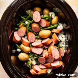 Sliced sausage, baby potatoes, green beans, and chopped onions in a slow cooker, ready to be cooked.
