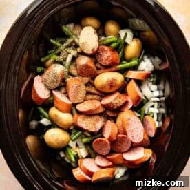 Sliced sausage, baby potatoes, green beans, and chopped onions with seasoning inside a slow cooker, ready to be cooked.