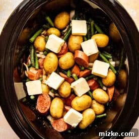 A slow cooker filled with whole baby potatoes, sliced sausage, green beans, onion pieces, and cubes of butter, ready to be cooked.