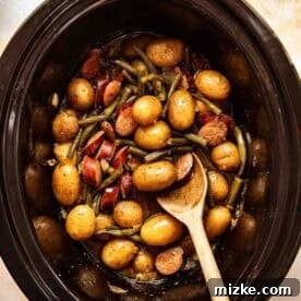 A slow cooker filled with small potatoes, green beans, sausage slices, and a wooden spoon.