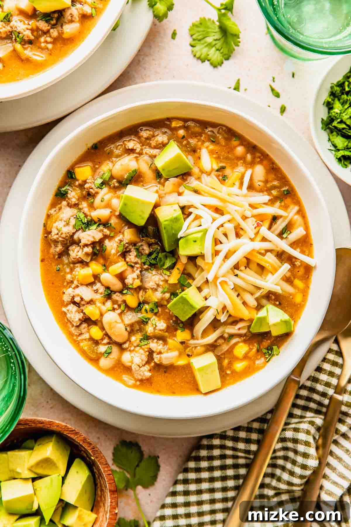 A vibrant bowl of ground turkey white bean chili, generously topped with shredded cheese and fresh diced avocado, perfectly garnished with cilantro. Nearby are small bowls of additional cilantro and avocado cubes, alongside a fork and spoon, inviting you to dig in.