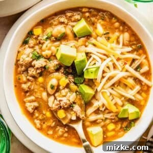 A close-up shot of a steaming bowl of white chili, brimming with ground turkey, corn, and white beans. It's garnished beautifully with a sprinkle of shredded cheese, fresh avocado cubes, and green herbs, with a spoon resting in the bowl, inviting you to take a bite.