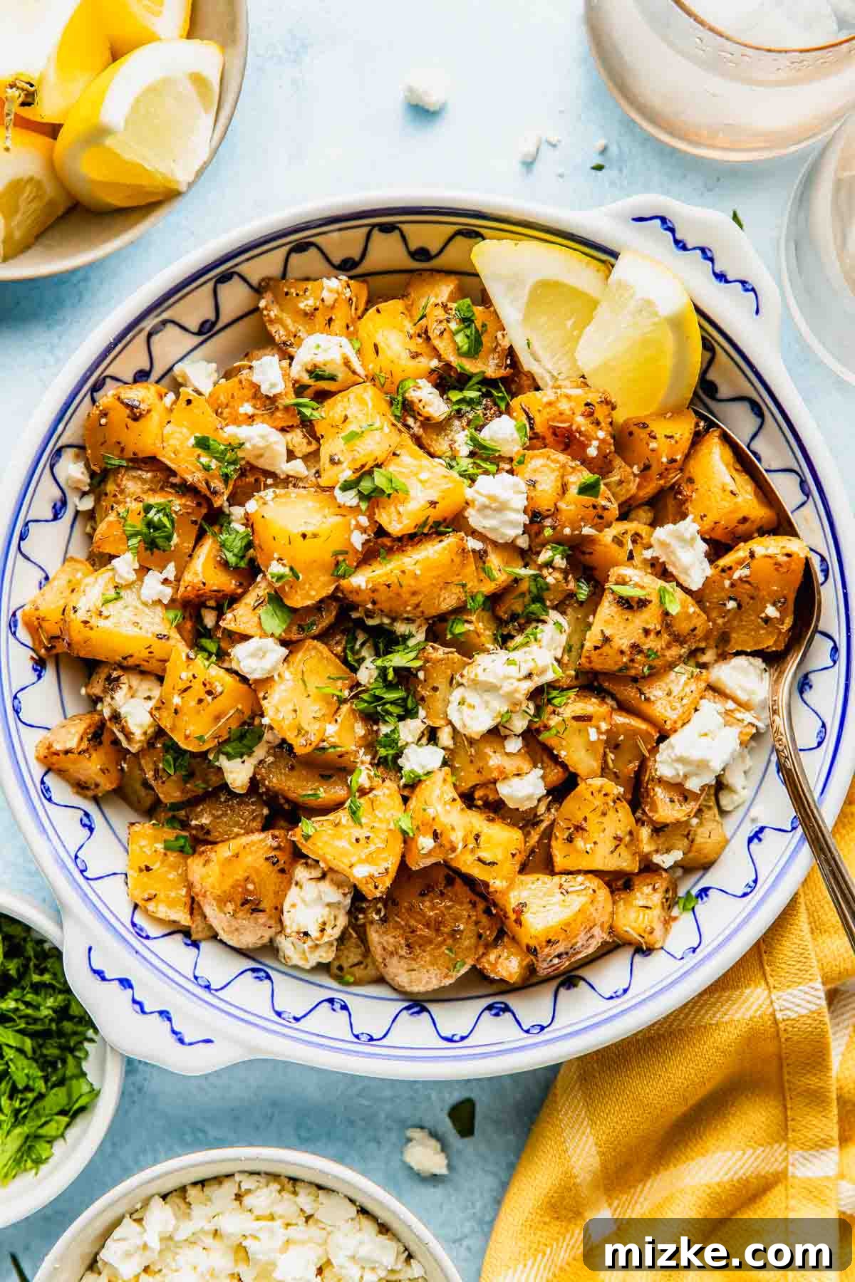 Zesty Greek Lemon Roasted Potatoes 2 A bowl of roasted Greek lemon potatoes, generously topped with crumbled feta cheese, freshly chopped herbs, and vibrant lemon wedges, with a serving spoon and extra garnishes nearby.