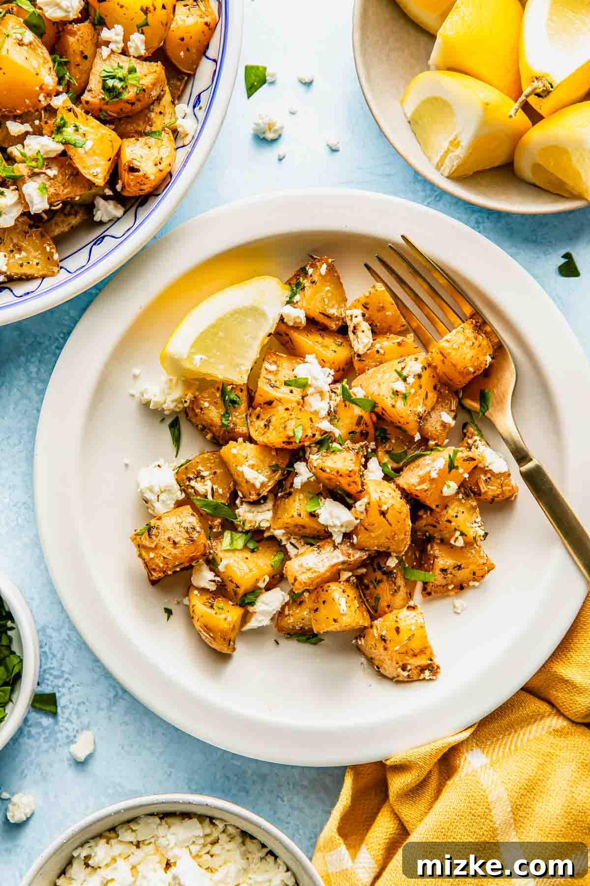 Zesty Greek Lemon Roasted Potatoes 3 A beautifully arranged plate of roasted Greek lemon potatoes, elegantly garnished with crumbled feta cheese and fresh herbs, served alongside a vibrant lemon wedge and a fork. Additional potatoes, lemon slices, and cheese are artfully placed nearby, creating a inviting culinary scene.