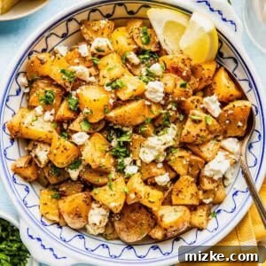 A bowl of roasted potatoes topped with crumbled feta cheese, chopped herbs, and two lemon wedges, with a spoon and garnishes nearby.