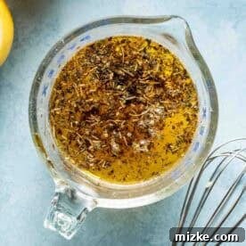 A glass measuring cup with a herb and oil mixture next to a metal whisk and sliced lemons on a blue surface.