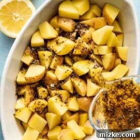 Cubed potatoes in a white baking dish are topped with herbs and seasoning, with a mixture being poured over them. Two halved lemons are beside the dish.