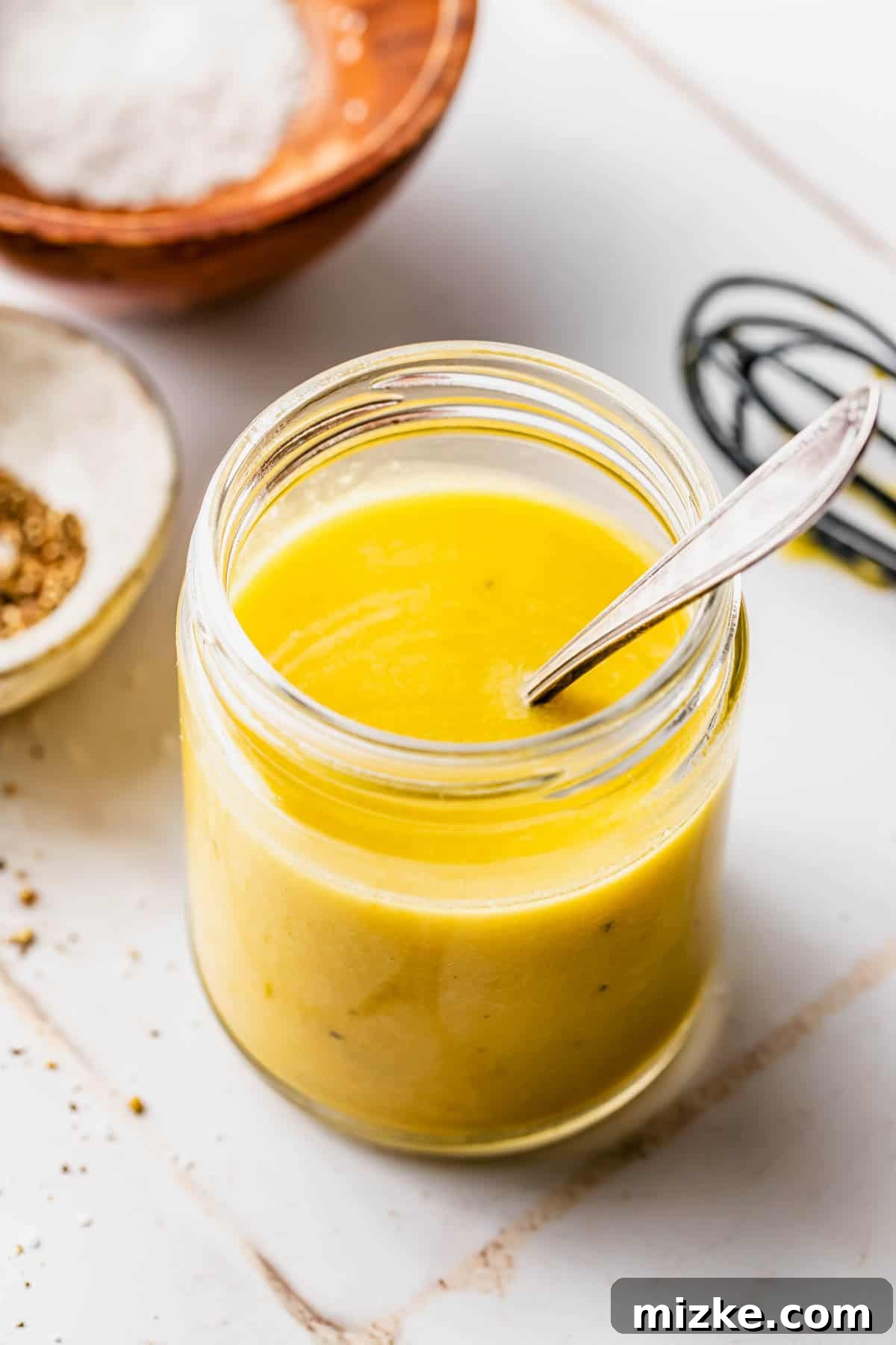 A clear glass jar filled with golden yellow Dijon vinaigrette, with a small metal whisk resting inside. In the blurred background, small bowls of sea salt and black peppercorns add to the rustic kitchen setting.