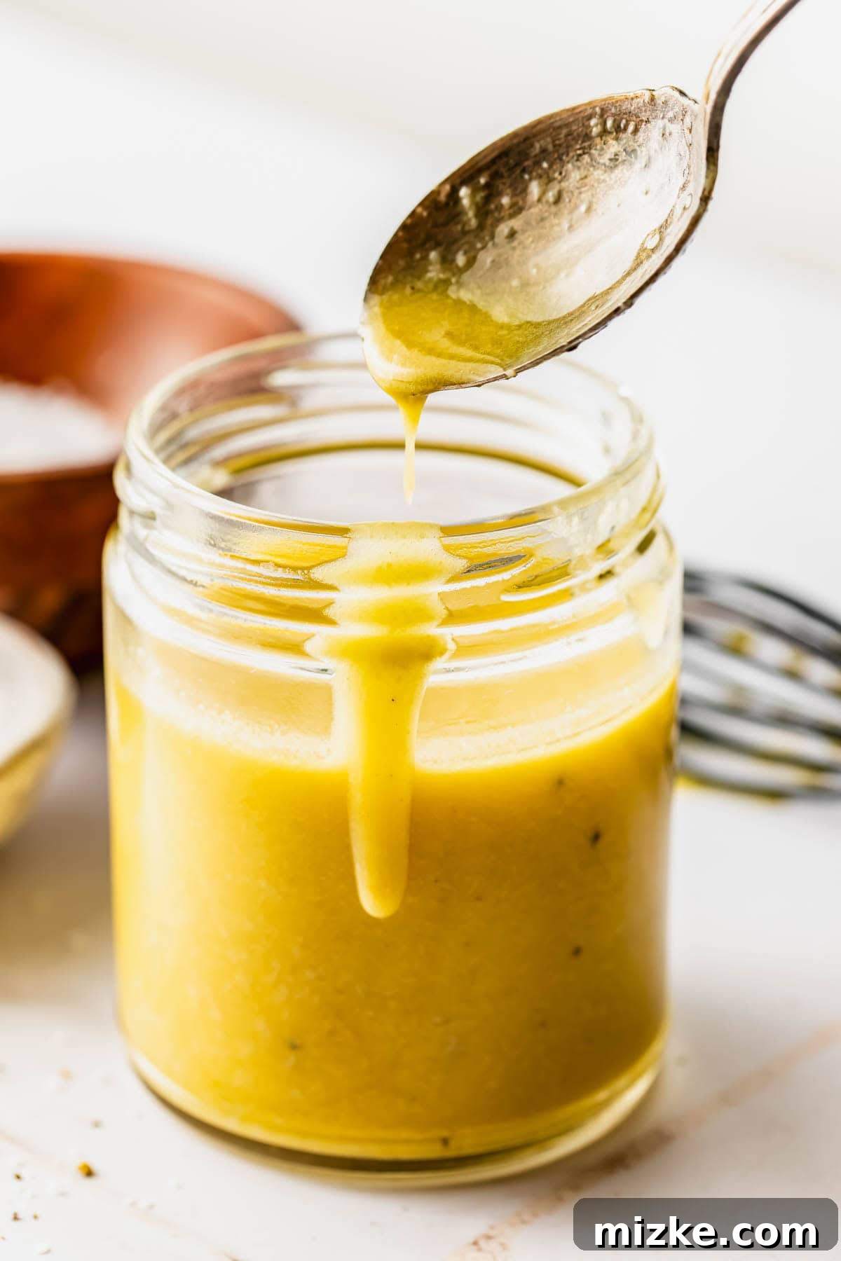 A stream of golden Dijon vinaigrette being poured from a silver spoon into a glass mason jar. A metal whisk and a wooden bowl are subtly visible in the soft-focus background, all set on a clean white surface.