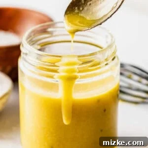 A spoon drips golden Dijon vinaigrette into a glass jar, with a metal whisk and a wooden bowl in the background on a white surface.