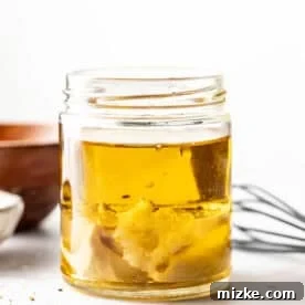 A glass jar containing olive oil and Dijon mustard, with a metal whisk and wooden bowl in the background on a white surface, illustrating the initial step of mixing.