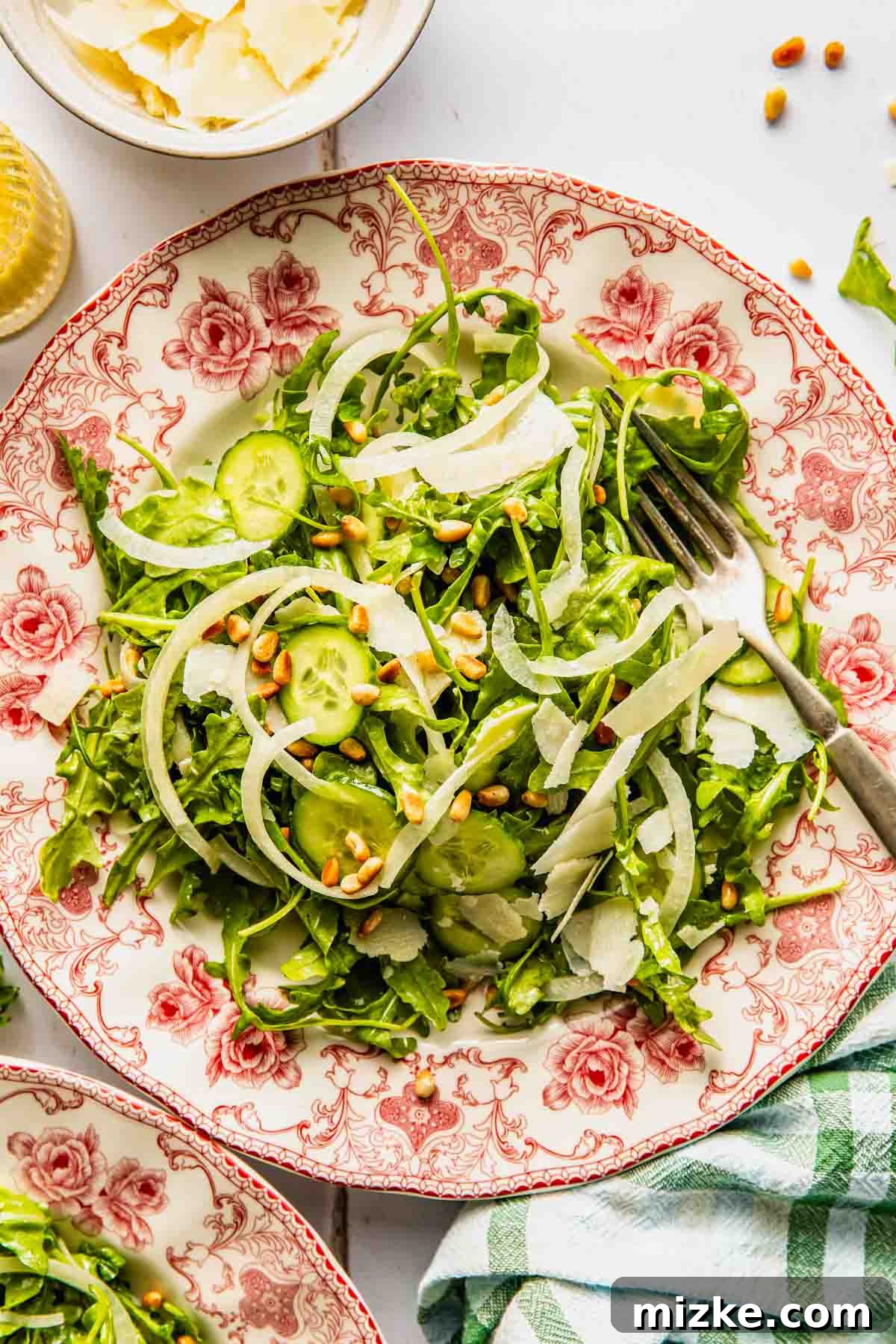 Peppery Rocket Greens 2 A vibrant simple arugula salad with sliced cucumbers, onions, shaved cheese, and pine nuts, served on a decorative plate with a fork.