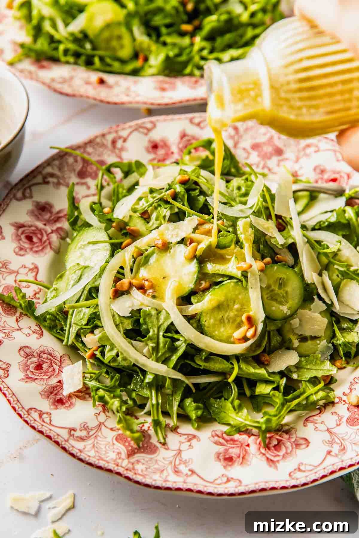 Peppery Rocket Greens 3 A hand drizzles homemade Dijon vinaigrette over a simple arugula salad with cucumber, shaved Parmesan, and pine nuts on a floral-patterned plate.