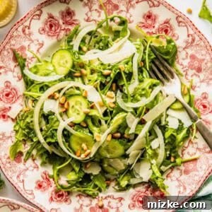 A salad with arugula, sliced cucumbers, onions, shaved cheese, and pine nuts is served on a decorative plate with a fork on the side.
