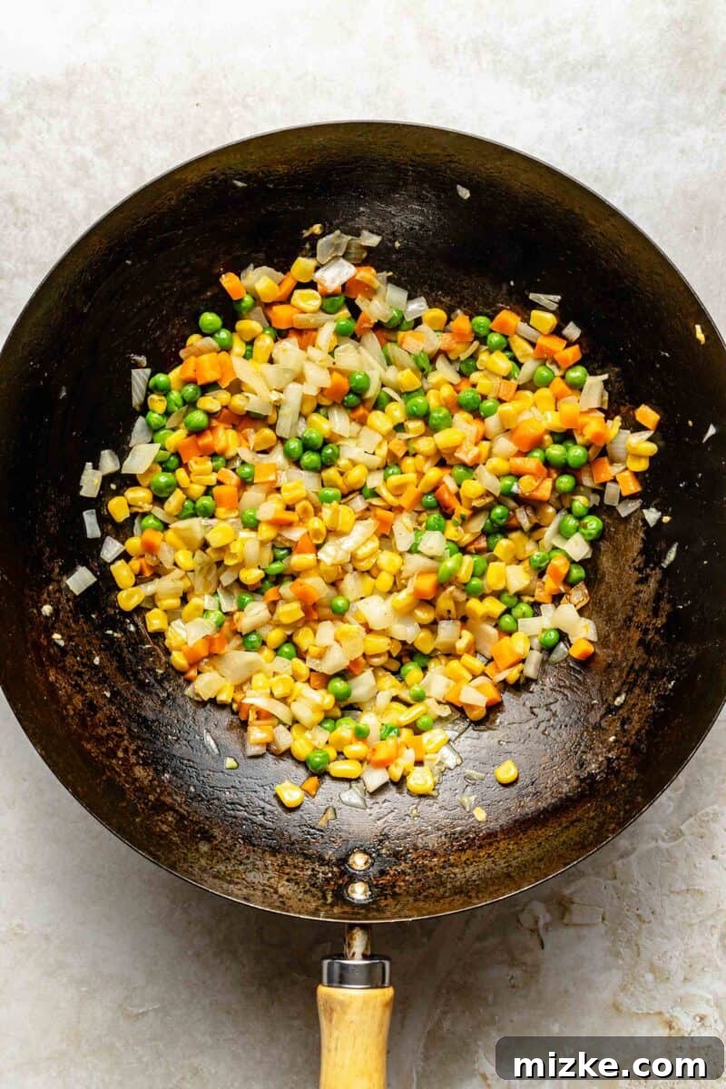 Sautéing vegetables until tender in a wok. Diced onions, corn, and peas with carrots mix being sautéed in a wok, vibrant and tender.