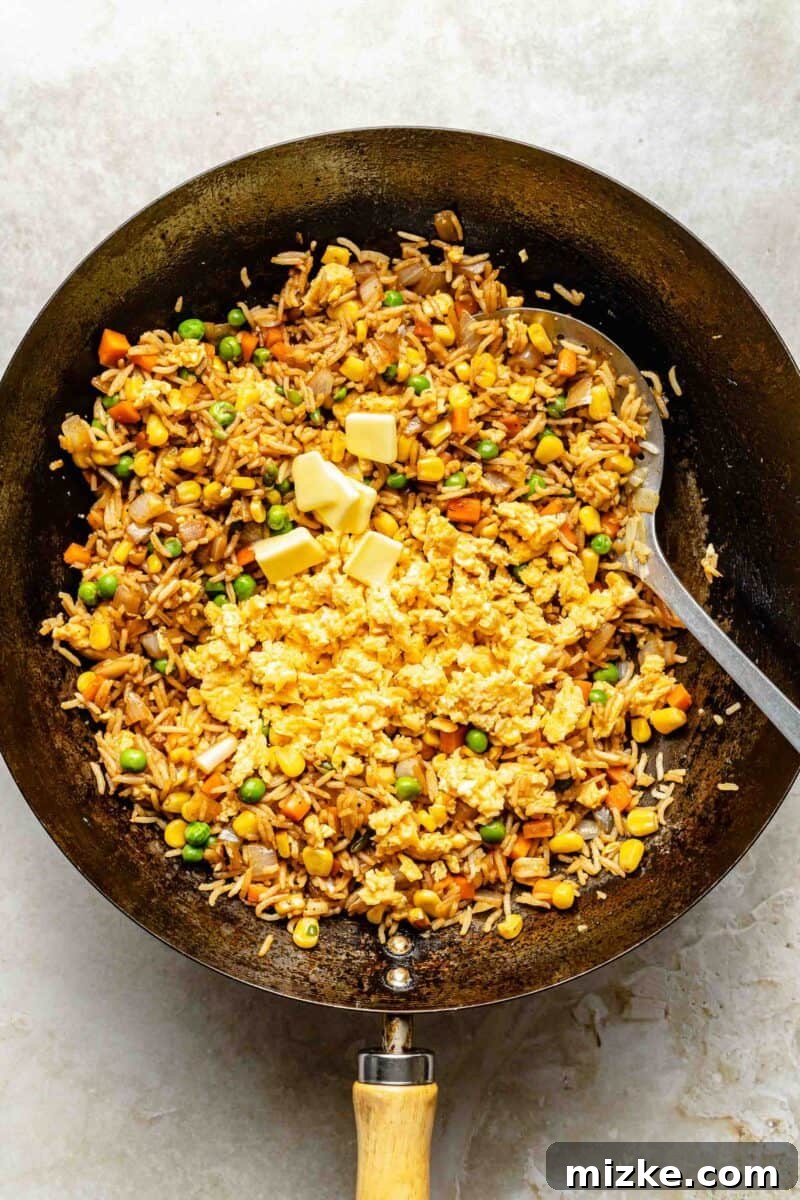 Folding scrambled eggs and butter into the fried rice. Cooked scrambled eggs and butter being stirred into the fried rice mixture in a wok, combining all ingredients.