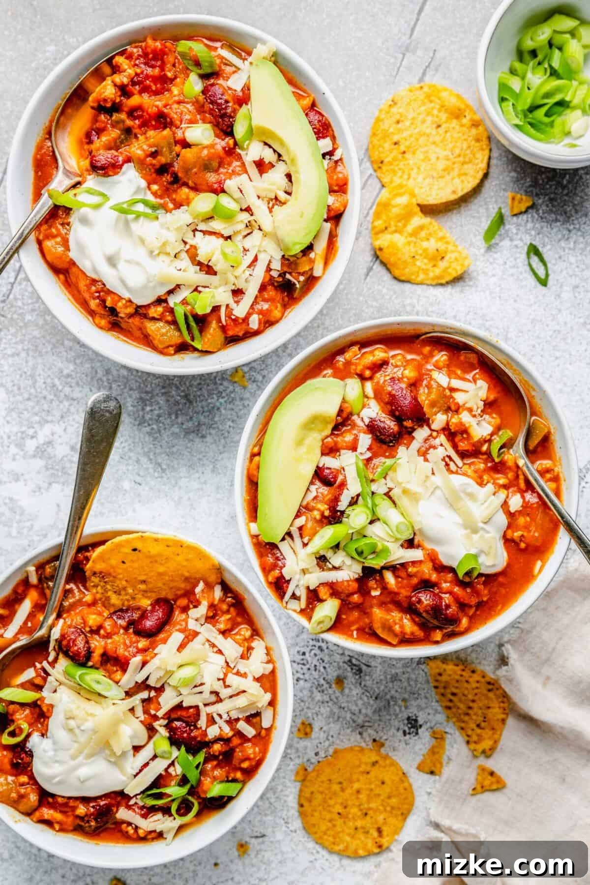 Three bowls of hearty turkey pumpkin chili garnished with fresh green onions, creamy sour cream, shredded cheese, crispy tortilla chips, and sliced avocado, each with a spoon ready for digging in.