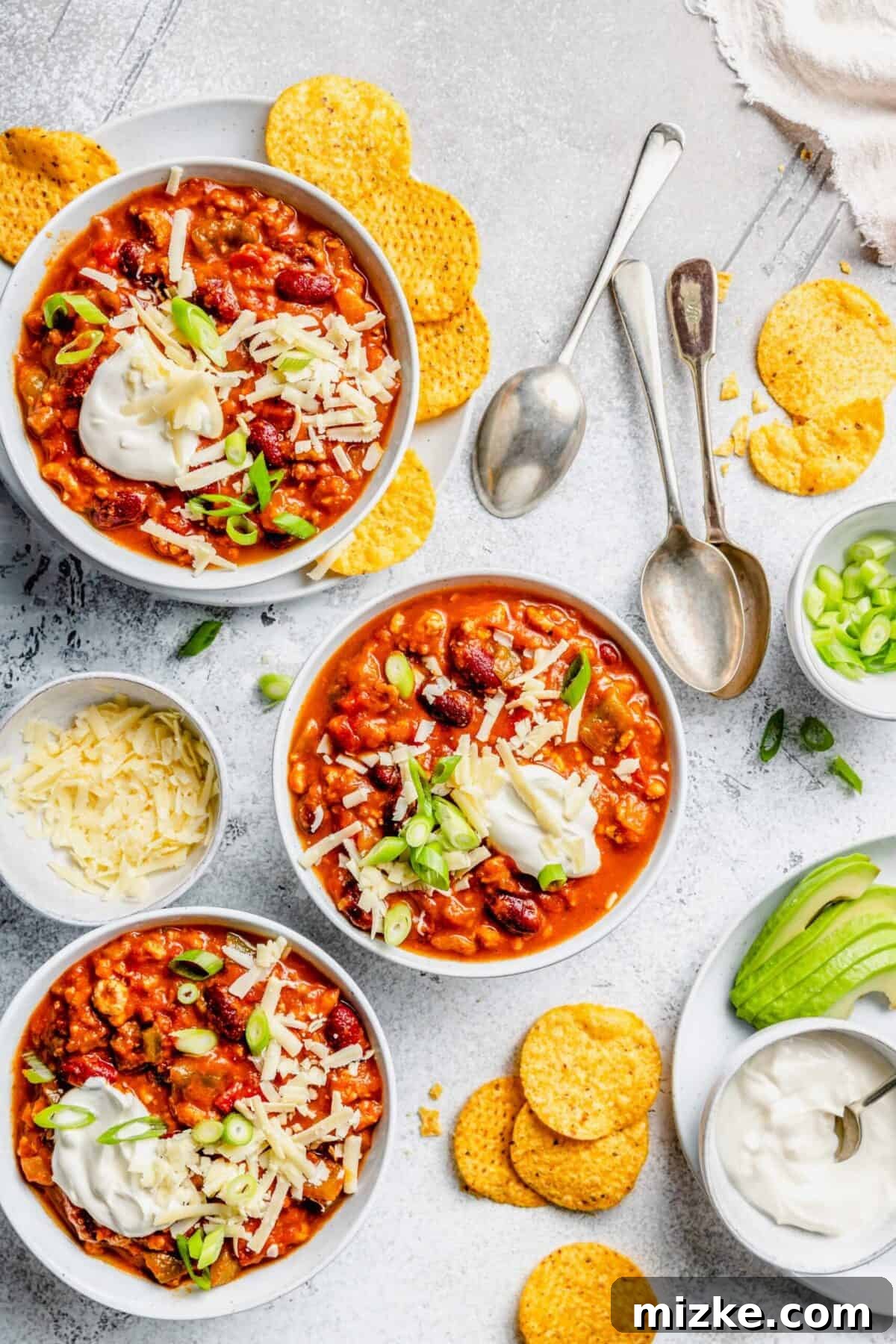 A beautifully composed shot of three bowls of Crockpot Turkey Pumpkin Chili, garnished with a generous sprinkle of shredded cheese, a dollop of sour cream, and vibrant green scallions. The bowls are set on a rustic table alongside fresh tortilla chips, a separate bowl of cheese, a serving of sour cream, and a plate of creamy sliced avocado, inviting everyone to customize their perfect bowl.