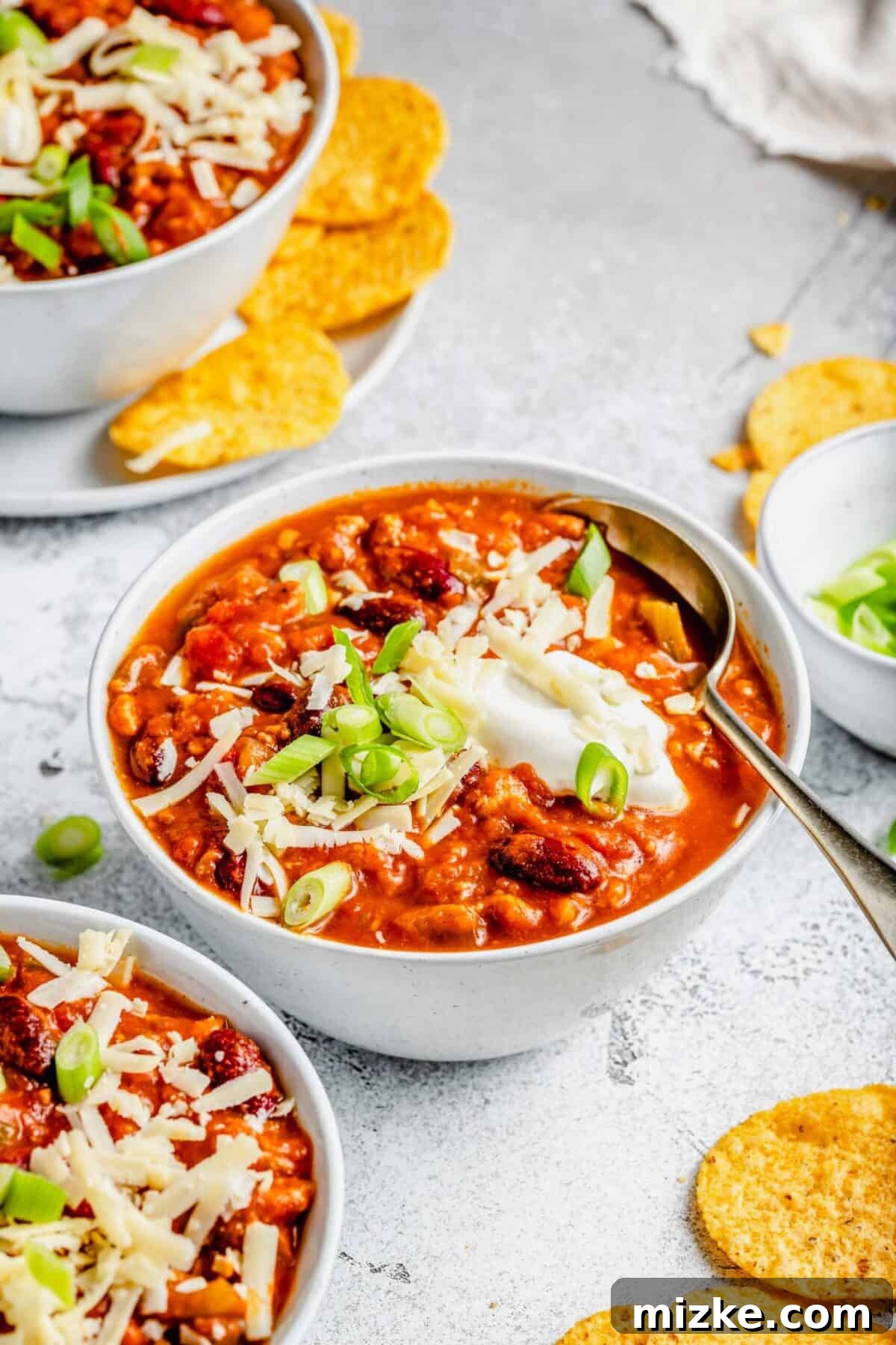 A close-up, inviting shot of a single bowl of Crockpot Turkey Pumpkin Chili, generously garnished with a swirl of sour cream, vibrant green scallions, and a sprinkle of shredded cheese. A spoon is nestled in the chili, beckoning for a taste. In the background, two other bowls and a scattering of crispy tortilla chips complete the cozy scene.