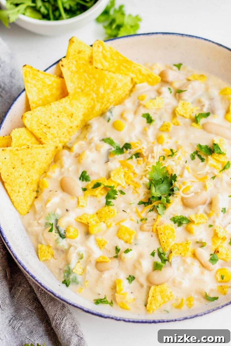White turkey chili is seen in a white speckled bowl with blue rim, served on a grey towel, with crushed tortilla chips and cilantro leaves as garnish.
