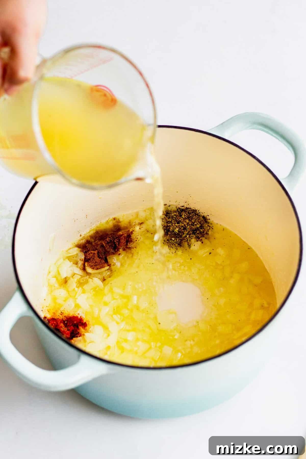 Pouring turkey stock into a large pot with cooked onions and garlic. Seasonings such as oregano, cumin, and cayenne can be seen sprinkled into the soup.