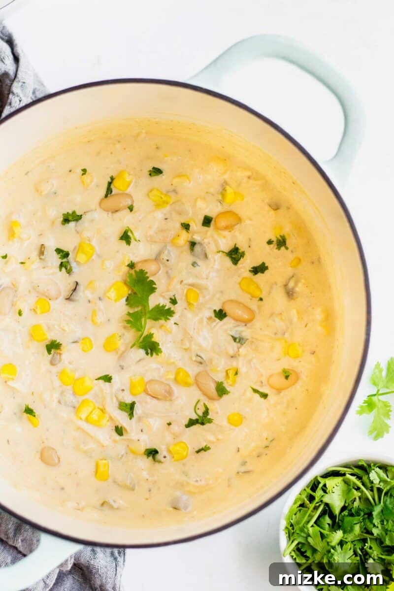 Overhead shot of large pot with white turkey chili garnished with a large sprig of cilantro leaf. Corn kernels and white beans can be seen dotting the soup throughout. Fresh cilantro leaves are on the bottom right corner and a greyish towel is underneath the pot.