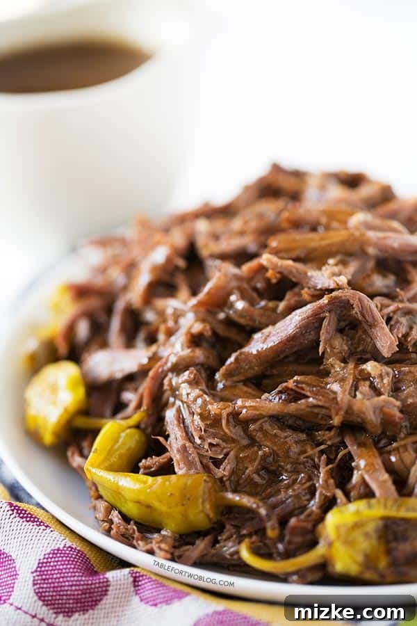 Experience effortless cooking and incredible flavor with Mississippi Roast – a 5-ingredient slow cooker recipe delivering tender beef and satisfying meals, perfect for dinner and delicious leftovers. A close-up of tender, shredded Mississippi Roast on a white plate, garnished with vibrant pepperoncini. The plate rests on a purple polka-dotted towel, with a gravy boat subtly blurred in the background, hinting at the rich flavors within.