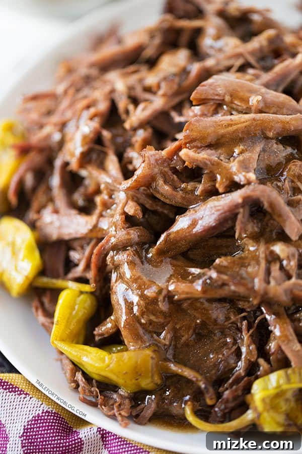 Sink your teeth into the ultimate comfort food: Mississippi Roast, slow-cooked to perfection with a rich, tangy sauce and tender beef. It's truly satisfying and packed with flavor! A tantalizing close-up of shredded Mississippi Roast, generously coated in its rich au jus, with bright green pepperoncini visible along the edge of a white plate. The succulent beef promises an explosion of flavor.