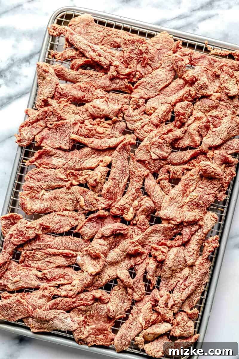 Preparing beef for optimal crispiness by coating and freezing. Thinly sliced beef strips coated in soy sauce and cornstarch, laid out on a wire rack over a baking sheet, ready for freezing.