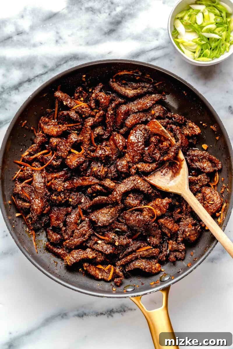 Tossing crispy beef in its vibrant, sweet, and spicy sauce. Freshly fried crispy beef strips coated in a glossy, orange-infused sauce in a large pan.