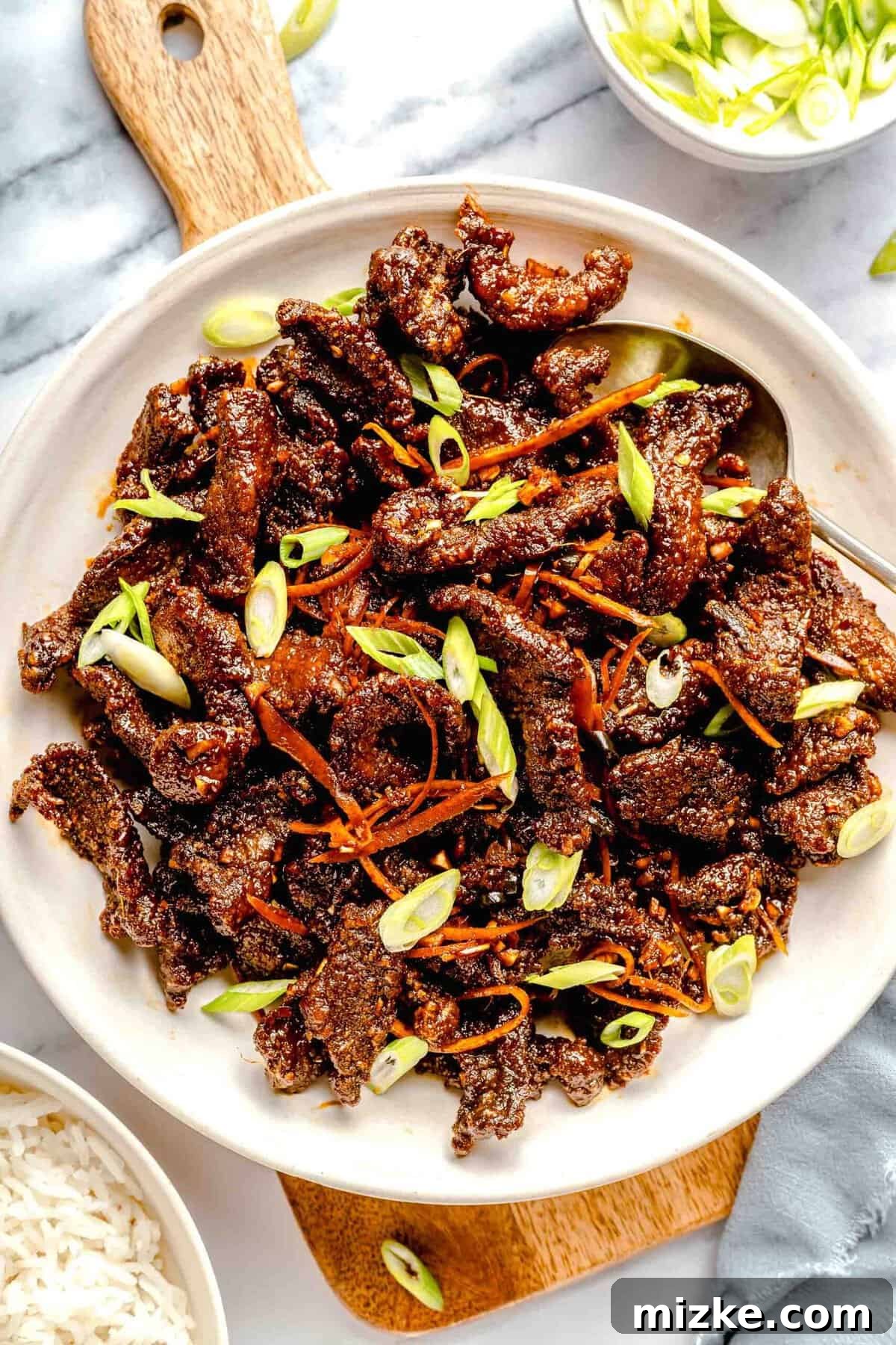 Crispy beef, freshly tossed in sauce and ready to be served. An overhead view of crispy beef on a large white serving platter, beautifully garnished with fresh green scallions.